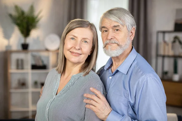 Oturma odasında dururken kameraya şefkatle ve sevgiyle bakan son sınıf evli çiftin portresi. Sevgili koca, karısının omzuna nazikçe dokunuyor. Aile hazinesi kavramı.