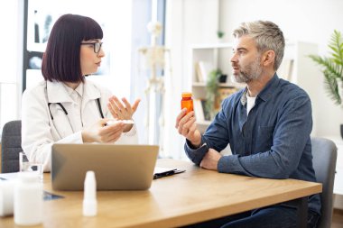 Beyaz üniformalı kadın pratisyen. Yeni klinikte elinde ilaç şişesi olan olgun bir hastayla iletişim kuruyor. Doktor sağlık sorunlarını tartışıyor ve laptoptaki tıbbi verileri inceliyor..