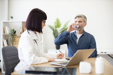 Gri saçlı hasta yeni klinikteki pratisyen hekimle randevusunda bir bardak soğuk su içiyor. Genç profesyonel reçete yazıyor ve müşterilerle çalışmak için uzaktan bilgisayar kullanıyor.