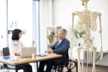 Genç hekim hasta kontrolü yaparken iskelet modelinin doktor muayenehanesine yerleştirilmesine odaklanın. Hastanedeki erkek tekerlekli sandalye kullanıcısının tıbbi durumunu teşhis eden ortopedist..