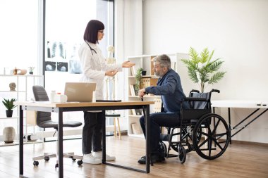 Laboratuvar önlüklü genç bir kadın, bastonlu olgun bir adama danışma odasında tekerlekli sandalyeden kalkması için yardım ediyor. Yaraları iyileşme sürecinde hastanın dengesini bulmasına yardımcı olan etkili bir terapist.