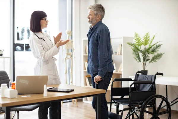 stock image Smiling female with stethoscope clapping while happy man standing on his feet after using wheelchair. Cheerful doctor in consulting room congratulating patient with regaining mobility after accident.