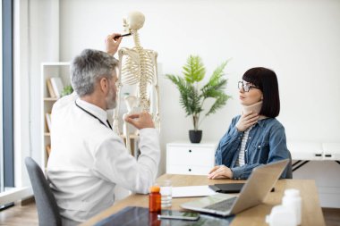 Çekici kadın doktor masasında otururken beyaz önlüklü kendine güvenen adam iskeletin omurgasını işaret ediyor. Ensesinden yaralanan genç bir kadın aile hekimini klinikte ziyaret ediyor..