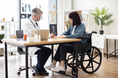 Gözlüklü güzel bir kadın hastanede yazı masasında otururken doktor ceketinde tablet tutan kendine güvenen bir yetişkin. Verimli travma uzmanı hastayla tomografi görüntülerini analiz ediyor..