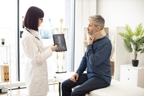 stock image Caucasian lady holding tablet with CT scan image on screen while talking to male in neck brace sitting on exam couch. Young orthopedist analyzing thoracic vertebrae on digital report in clinic.