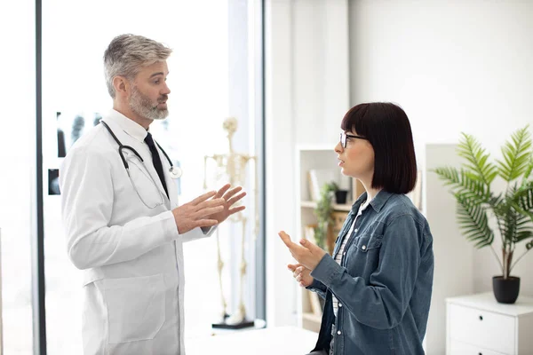 迷人的年轻女人手牵着喉咙向穿着实验室外套的留胡子男人描述健康状况 在现代医院为女性病人提供有关疾病的咨询的自信治疗师 — 图库照片