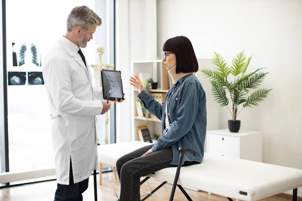 stock image Caucasian woman in neck brace looking at x-ray image on digital device held by bearded therapist in lab coat. Medical specialist explaining test results to patient with walking cane in exam room.