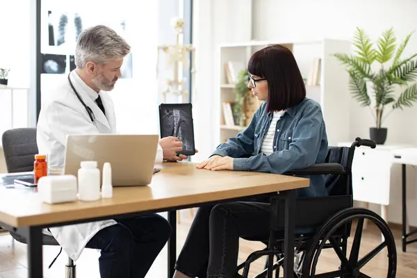 stock image Confident adult in doctors coat holding tablet while pretty lady in glasses sitting in wheelchair at writing desk in hospital. Efficient traumathologist analyzing CT scan image with patient at work.