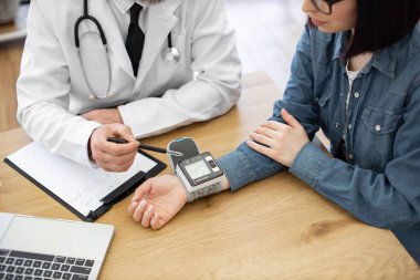 Gözlüklü kadının beyaz önlüklü doktorla masada otururken bilek tipi cihaz taktığı görüntüyü kapat. Genel pratisyen doktor hasta kaydına kaydetmeden önce ölçümleri tahmin ediyor.