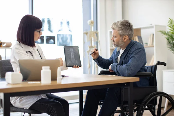 stock image Attractive caucasian man taking pen for marking on tablet with x-ray image carried by radiologist in consulting room. Confident specialist discussing results of diagnostic tests with wheelchair user.