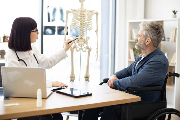 stock image Pretty woman in doctors coat pointing at human skeleton while mature male sitting in wheelchair at desk in consulting room. Intent therapist illustrating site of spinal injury on medical model.