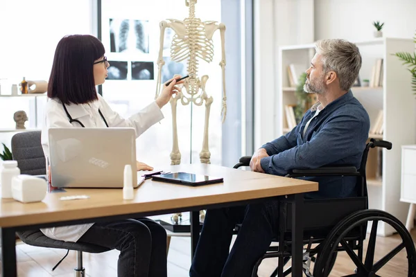 stock image Pretty woman in doctors coat pointing at human skeleton while mature male sitting in wheelchair at desk in consulting room. Intent therapist illustrating site of spinal injury on medical model.