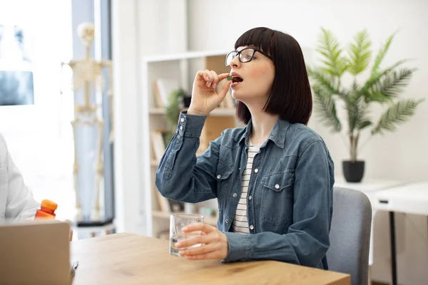 stock image Attractive woman in everyday wear taking round pill with glass of water while sitting at office desk in modern office. Charming brunette in spectacles being prescribed antibiotics by family doctor.