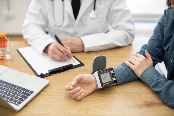 stock image Cropped view of womans wrist with electronic tonometer showing numbers of blood pressure. Male therapist writing numbers on paper clipboard for regulating treatment of illness at clinic.