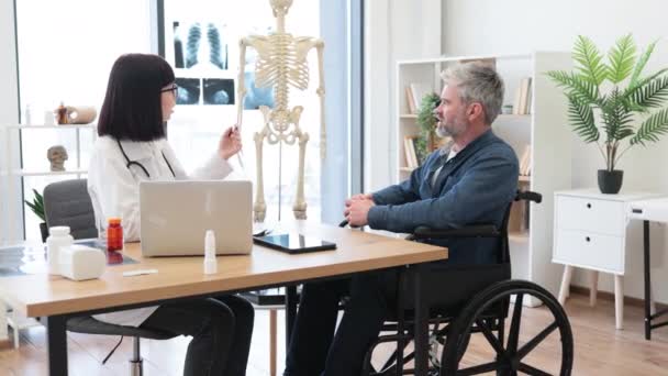 Брюнетка Халате Разговаривает Бородатым Мужчиной Инвалидном Кресле Сидя Столом Консультационной — стоковое видео