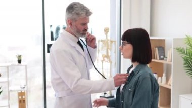 Kafkasyalı genç bir kadın nefes alırken soluk alıp verirken olgun bir erkek göğsünde steteskop tutuyor. Verimli bir pratisyen doktor, doktorların dinleme cihazıyla dişinin ciğerlerini kontrol ediyor..