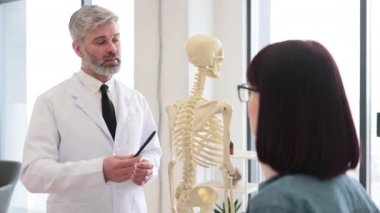 Omuz hizasında, boyunluk takan ve olgun bir adamın kullandığı insan iskeletine bakan güzel genç bir kadın. Gözlüklü endişeli bayan muayene için omurga zedelenmesinden sonra doktoru ziyaret ediyor..