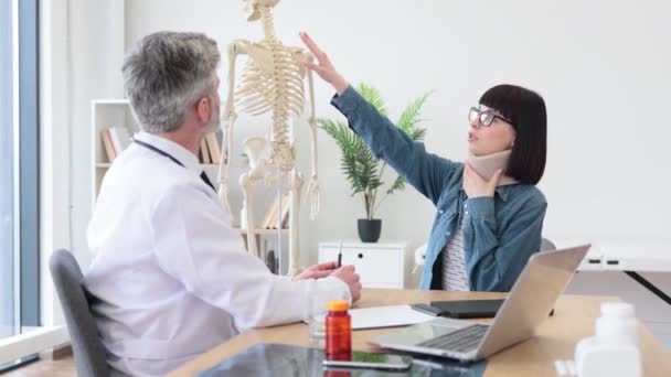迷人的女人坐在医生桌前 而有自信的男人穿着白衣 指着骨骼的颈椎 因颈部受伤而在诊所预约求医的年轻女性 — 图库视频影像