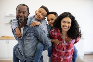 Close up portrait of multiethnic married couple with smiling kids on their backs practising artistic talents at home. Playful adults and children building new experiences together in room interior. clipart