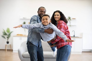 Aktif Afro-Amerikan yetişkinler evde hayali uçuşu test ederken mutlu çocuğu kollarında tutuyorlar. Kot pantolonlu enerjik çocuk anne ve babalar sayesinde özgürlük ve refah hissi yaşıyor..