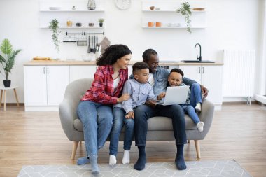 Gri kanepede yan yana otururken bilgisayar ekranına bakan çok ırklı yetişkinlerin ve çocukların tam boy görüntüsü. Tutkulu aile üyeleri çevrimiçi televizyon programının favori yarışmacısı için tezahürat yapıyorlar.