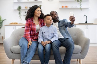 Kotlu giysiler içinde gülümseyen çok kültürlü bir aile güneşli bir günde oturma odasında otururken cep telefonu kamerasına bakıyor. Anaokulundaki oğluyla mutlu bir karı-koca büyükbabadan uygulamayla görüntülü arama alıyor..