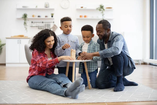 Friendly multiethnic family gathering together around coffee table on room floor at noon. Happy kids and involved parents building high tower of wooden blocks on floor of kitchen.