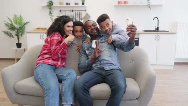 笑着多文化的父亲 电话和母亲显示V标志 而精力充沛的孩子站在沙发后面的家的休息厅 一对快乐的夫妇 他们的儿子在闲暇时通过手机应用程序获得了自画像 — 图库视频影像