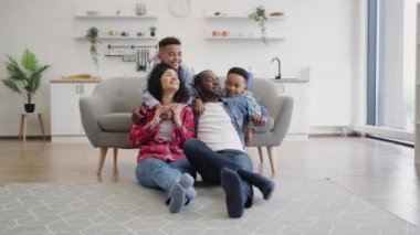 Küçük oğullar anne ve babaya arkadan sarılırken Afro-Amerikan eşlerin mutfak zemininde oturması. Dört kişilik sevgi dolu aile hafta sonları evde boş zamanlarında eğleniyor..