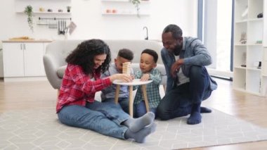 Çok yönlü gülümseyen yetişkinlerin ve anaokulu öğrencilerinin açık-planlı mutfakta sehpanın etrafında kucaklaşmalarının tam boy portresi. Sevgi dolu ebeveynler ve sevimli oğullar yıkılmış kuleye taş koyarken mutlu hissediyorlar..