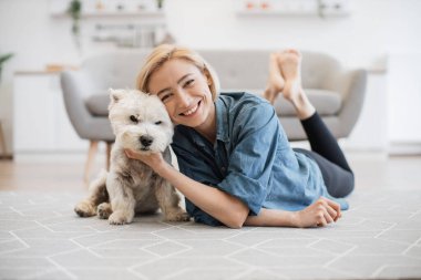 Kot gömlekli, sevimli gülümseyen bir kadının mutfak zemininde sevimli küçük bir Westie 'yi okşayan uzun bir portresi. Neşeli hayvan bakıcısı evde iyi huylu küçük bir hayvanla poz veriyor..