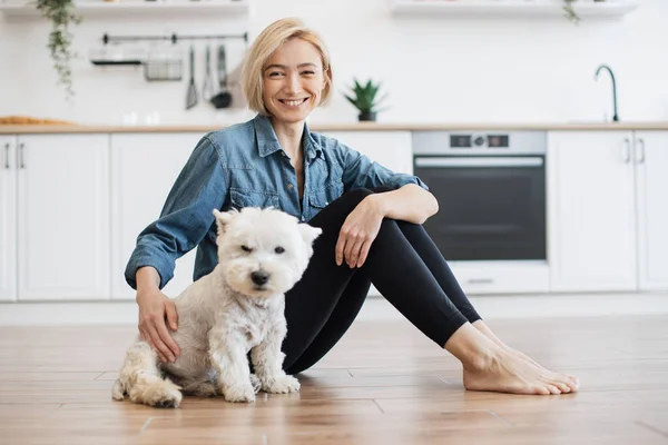Portrait of cheerful young lady sitting next to well-behaved white dog on bare floor in kitchen interior. Affectionate female keeper giving canine friend overall attention on bright sunny day.