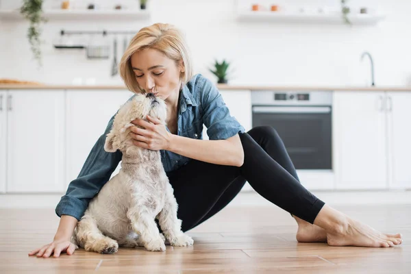 Friendly caucasian female kissing white terriers nose while resting on hard surface on kitchen background. Loving dog owner in casual wear displaying affection for furry buddy after training.