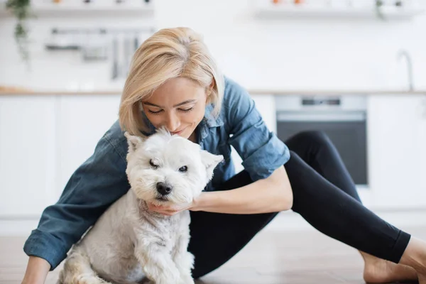 Friendly caucasian female kissing white terriers nose while resting on hard surface on kitchen background. Loving dog owner in casual wear displaying affection for furry buddy after training.