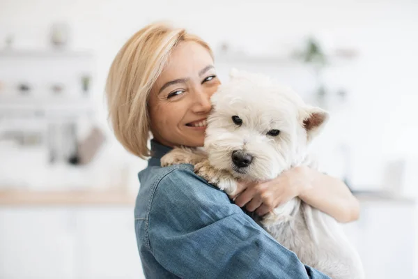 Portrait of smiling blonde lady with West Highland White Terrier in arms posing on background of spacious kitchen. Happy caucasian pet owner rewarding good behavior during day with gentle hug.