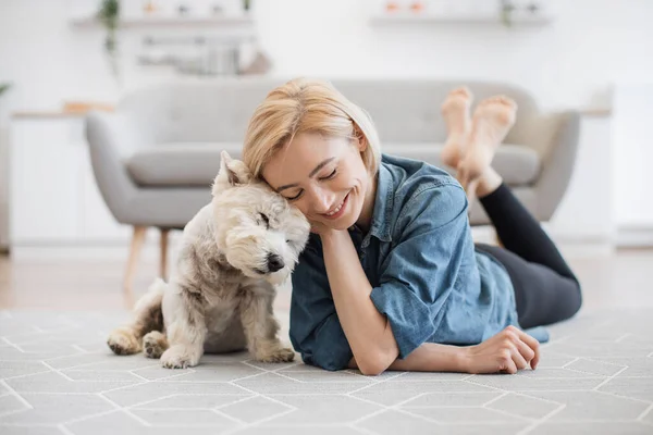 Kind female person and faithful small terrier snuggling to each other with heads while staying on wooden floor at home. Affectionate woman and cuddly Westie enjoying bonding interaction on sunny day.