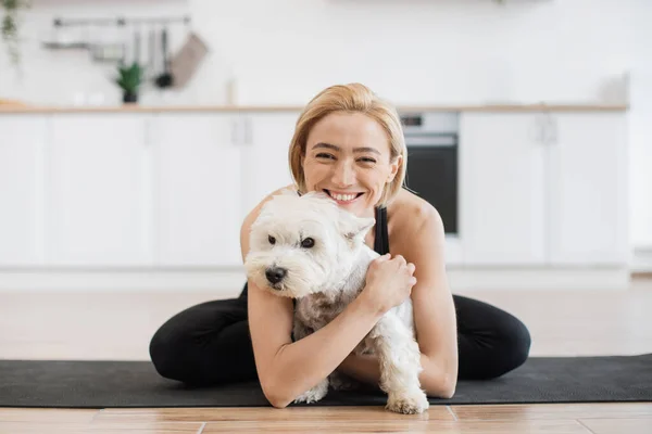 stock image Affectionate adult female in yoga activewear embracing white terrier while sitting cross-legged on rubber mat indoors. Healthy lady awarding sweet pet with hug for good behaviour during home workout.