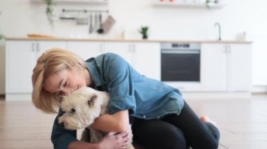 Gülen kadının portresi, tahta zeminde bacak bacak üstüne atmış şekilde poz verirken nazikçe sevimli köpeğe sarılıyor. Akşam yemeğinden sonra parka gitmeden önce sevgili evcil hayvanına iyi baktığını gösteren mutlu bir bayan..