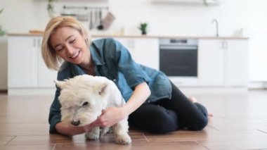 Yetişkin kadınlarda kalan sadık West Highland White Terrier modern bir dairede yaşarken kucaklaşıyor. Sevgi dolu evcil hayvan sahibi, tüylü en iyi arkadaşını öperken köpekle günlük etkileşiminden zevk alıyor..