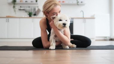 Yoga kıyafetleri içinde sevimli yetişkin bir kadın. Beyaz teriyeri kucaklıyor. Kapalı alanda lastik paspasın üzerinde bağdaş kurarak oturuyor. Sağlıklı bir bayan, ev ödevi sırasında iyi halden evcil hayvanı kucaklayarak ödüllendiriyor..