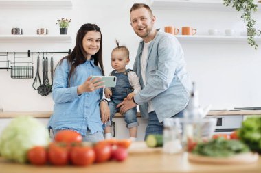 Akıllı telefonlu çekici bir kadın, dikkatli bir baba modern mutfakta taze ürünlerle yemek masasına bakarken. Sevgi dolu eşler ve küçük kız internetten yemek videoları izliyorlar..