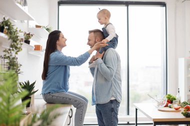 Mutfakta erkek omuzlarında oturan sevimli küçük bebeğe elini uzatan beyaz bir kadının yan görüntüsü. Neşeli eşler küçük bebekleri güvende tutuyorlar ve yemek hazırlama sürecinde izleniyorlar..