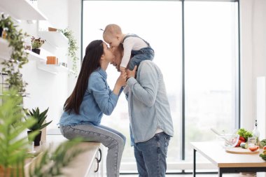 Sevimli anne, modern mutfağın atölyesinde otururken babasının omuzlarında tatlı bir bebekle burnuna dokunuyor. Sağlıklı beslenme için yemek hazırlayan küçük kızı da içeren dost canlısı ebeveynler..