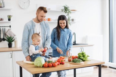 Kot gömlekli yakışıklı baba küçük kızına domates verirken güzel anne mutfak masasında bıçak ustalığı yapıyor. Aile aktivitelerinden zevk alırken küçük bebek öğrenme renkleri ve biçimleri.