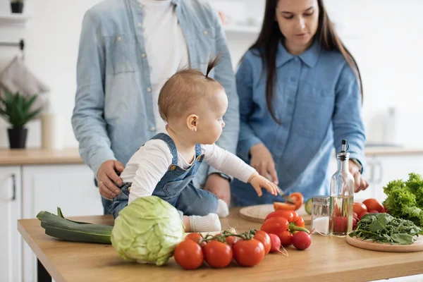 剪下黑发女子牵着黄瓜 而甜美的女婴伸出手去取绿色蔬菜的画面 可爱的年轻妈妈教女儿吃在家里一起准备的健康食品 — 图库照片