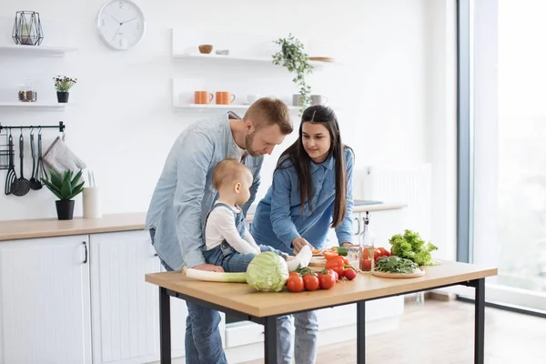 stock image Funny little girl in cozy outfit sitting on dining table while caring young mother holding bite-sized slice of tomato. Mindful parents motivating infant daughter to making healthier food choices.