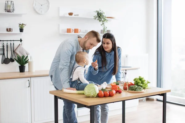 stock image Funny little girl in cozy outfit sitting on dining table while caring young mother holding bite-sized slice of tomato. Mindful parents motivating infant daughter to making healthier food choices.