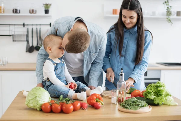 有爱心的爸爸吻着小女儿的额头 而可爱的小宝宝则坐在西红柿和蔬菜旁边的早餐桌上 在家里做饭时 敬重三个加强联系的家庭 — 图库照片