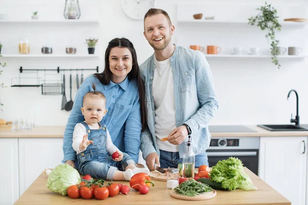 Portrait of happy three-person family in denim wear standing at kitchen table with assorted vegetables and greens. Stylish spouses achieving balanced diet by using organic products for home cooking.