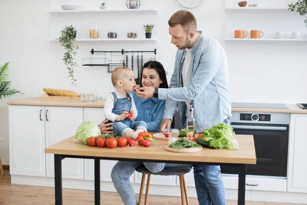 Mutlu bir aile babası, küçük kızını beslerken bir yandan da gülümseyen bir anne, mutfak sandalyesinde oturmuş, küçük kıza tezahürat yapıyor. Genç eşler tatlı domates ikram ederek çocuk diyetine yeni besinler ekliyorlar..